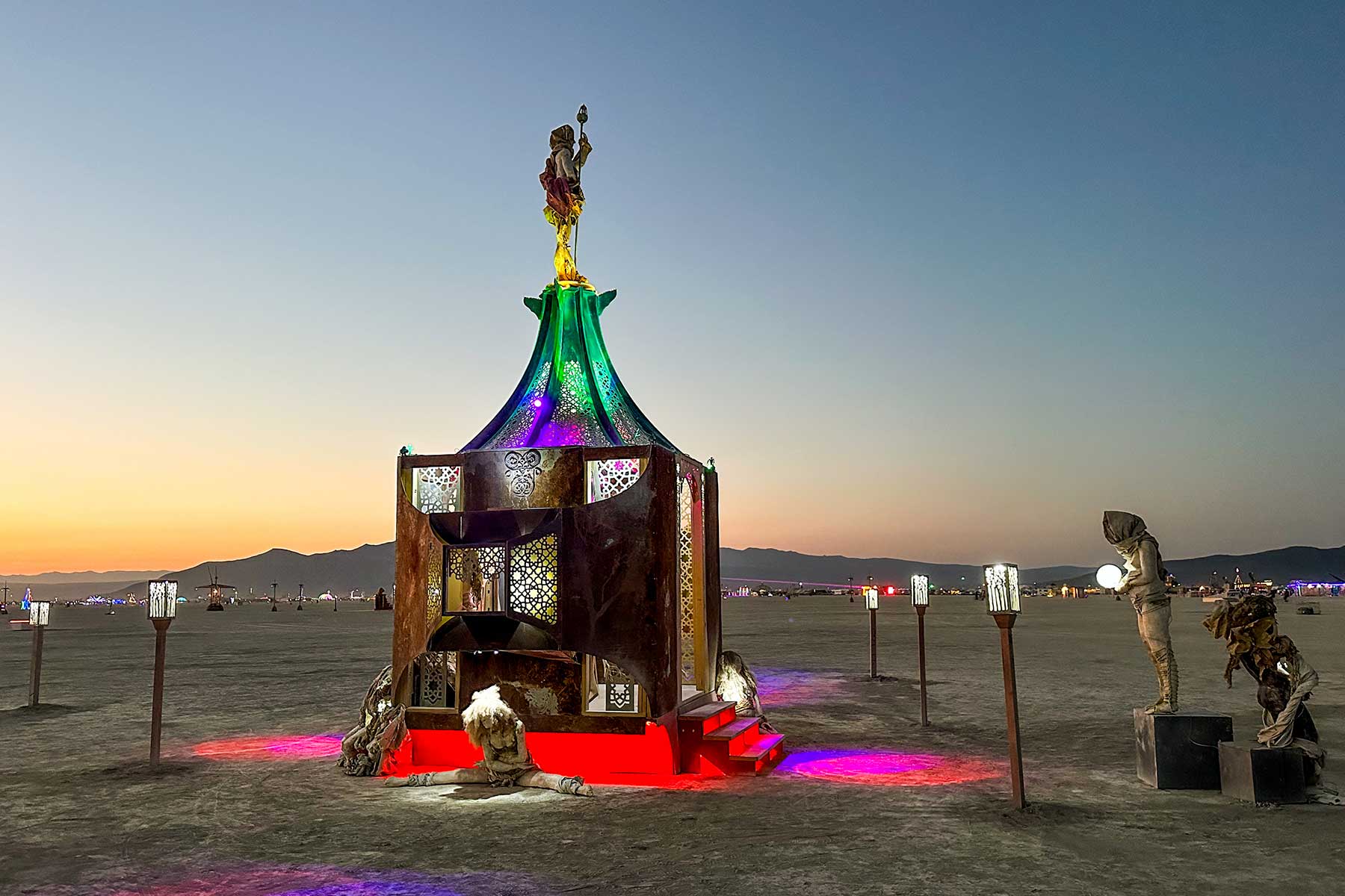 Playful Again at sunset: an ornate pavilion with a glowing rainbow canopy, topped by a dancing figure, above a pool of red light