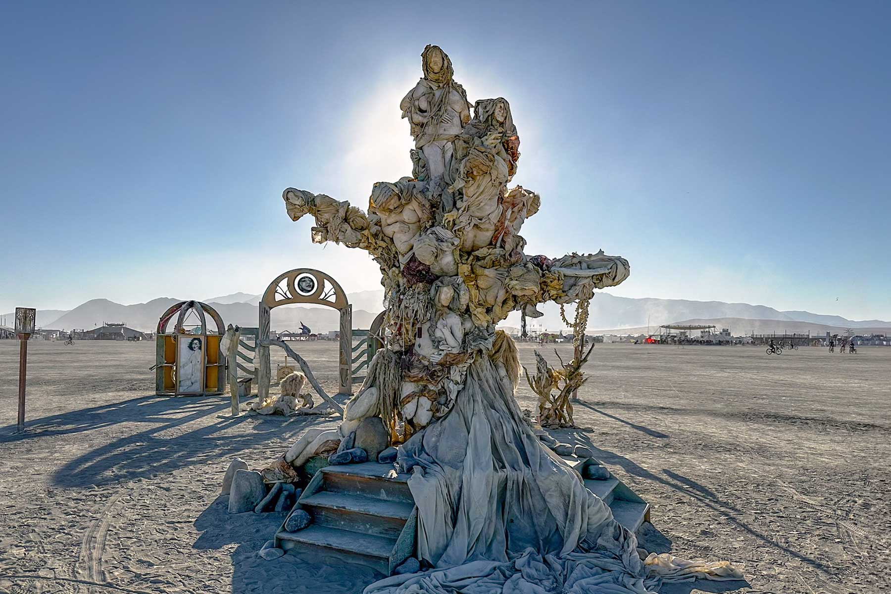 Womantree on the playa by day: a towering tree of intertwined cast figures rising from a draped base with steps, its arched entry gate behind it