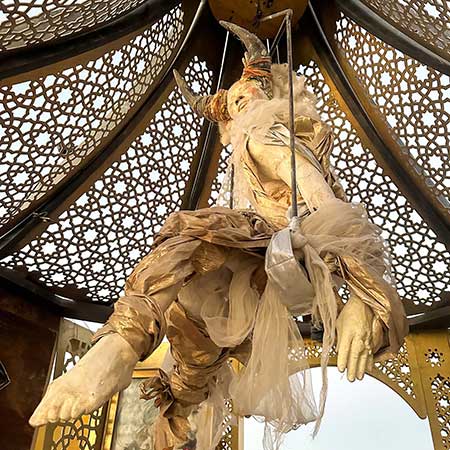 A cloth-draped figure suspended from the lattice dome inside the pavilion, seen from below