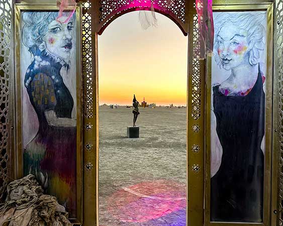 Painted portraits of two women framing the arched doorway, with a sculpture visible on the playa at sunset