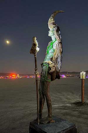 Night view of the horned figure with its fur-topped staff, lit green, with the moon and city lights behind