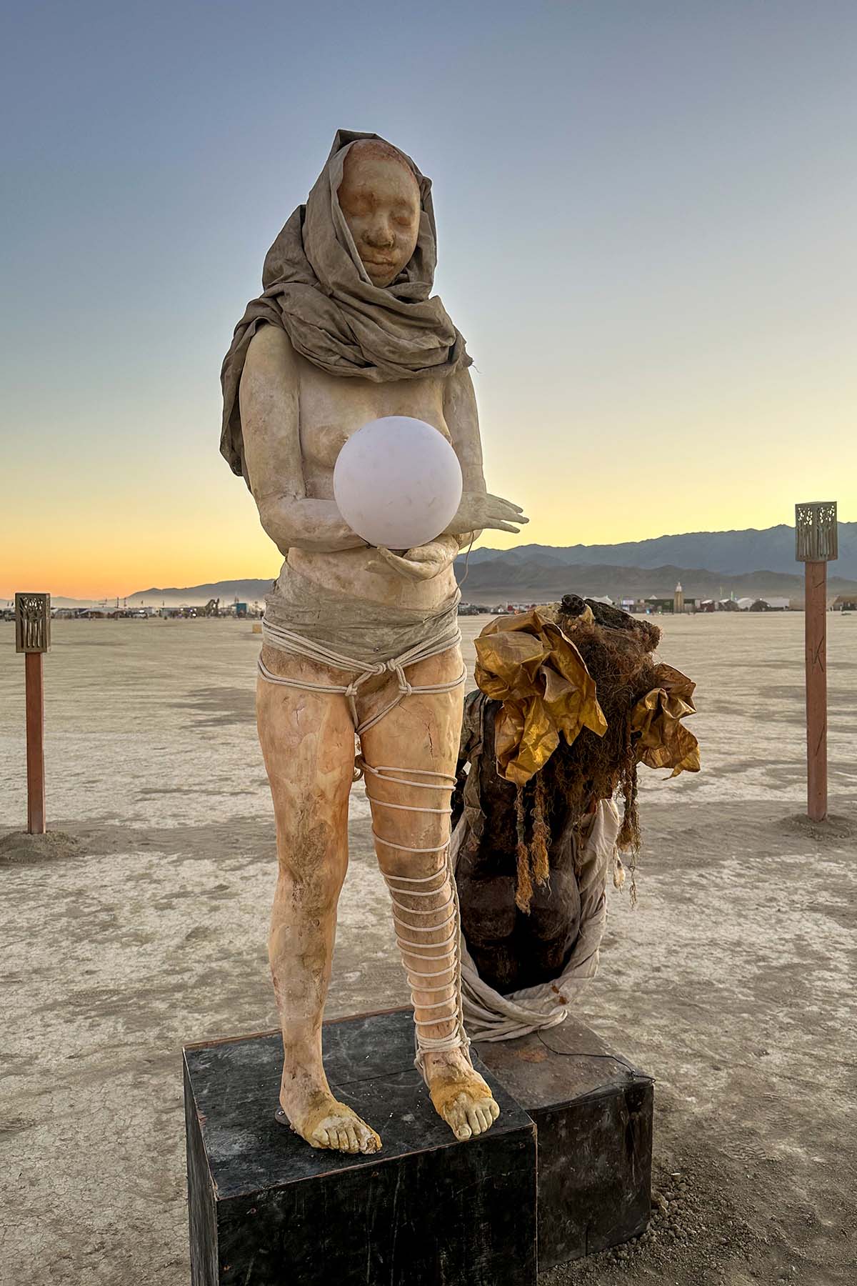 Hooded figure wrapped in cloth holding a glowing white orb, standing on a black plinth on the playa at sunset