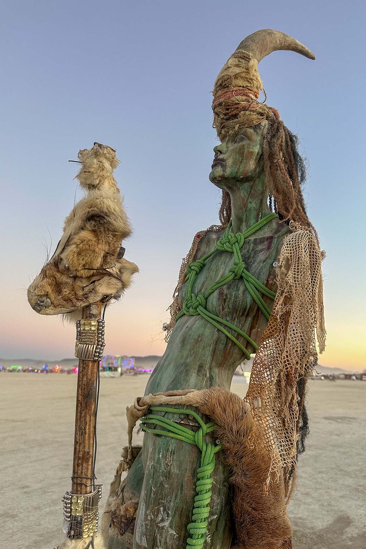 Close-up of a green-toned horned figure bound with green rope, holding a tall staff topped with fur and feathers