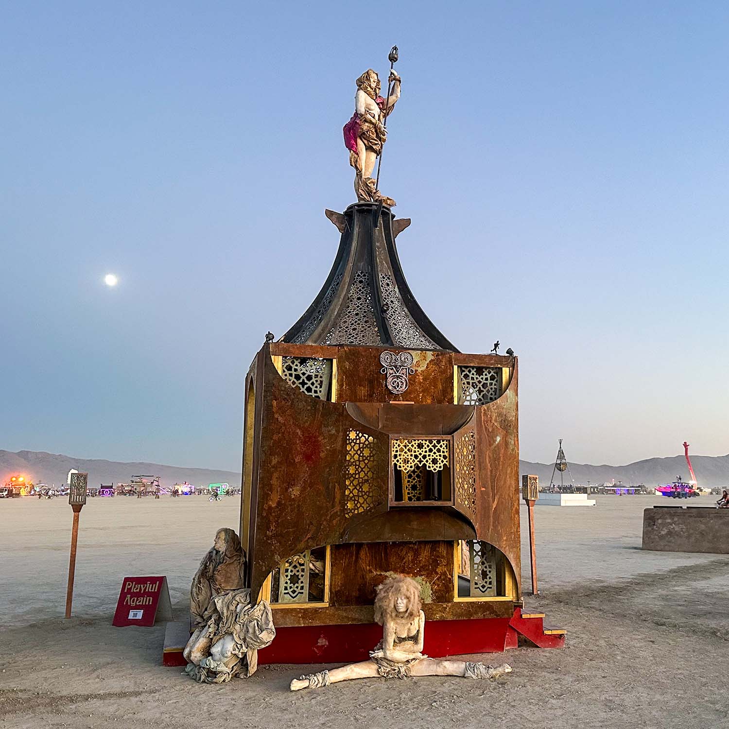 Front view of Playful Again at dusk, with the moon rising and sculptures kneeling and doing the splits at its base