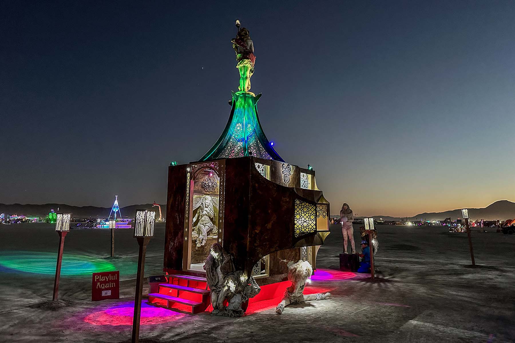 Playful Again at night, lit green, red and purple, with sculptures around the base and lights across the playa