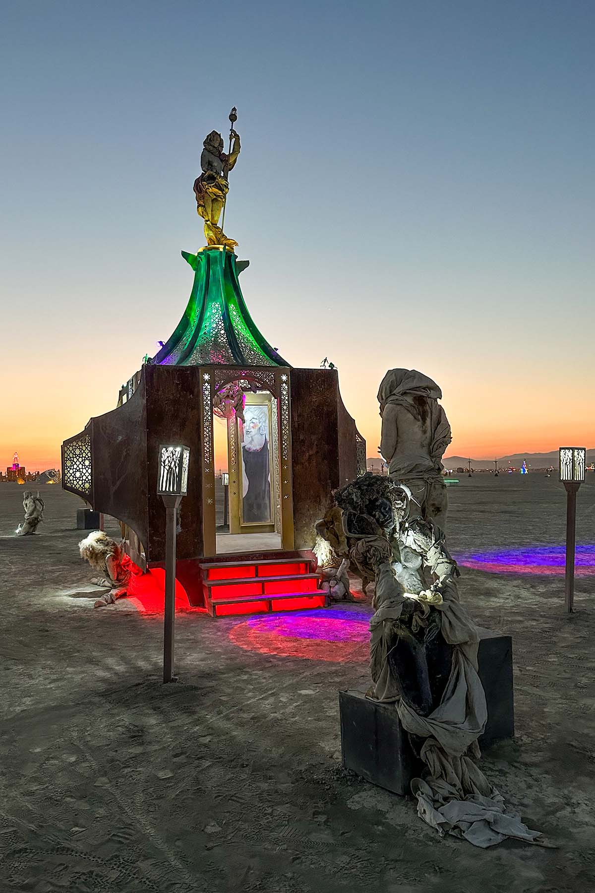 Draped sculptures on plinths surrounding the lit pavilion at sunset