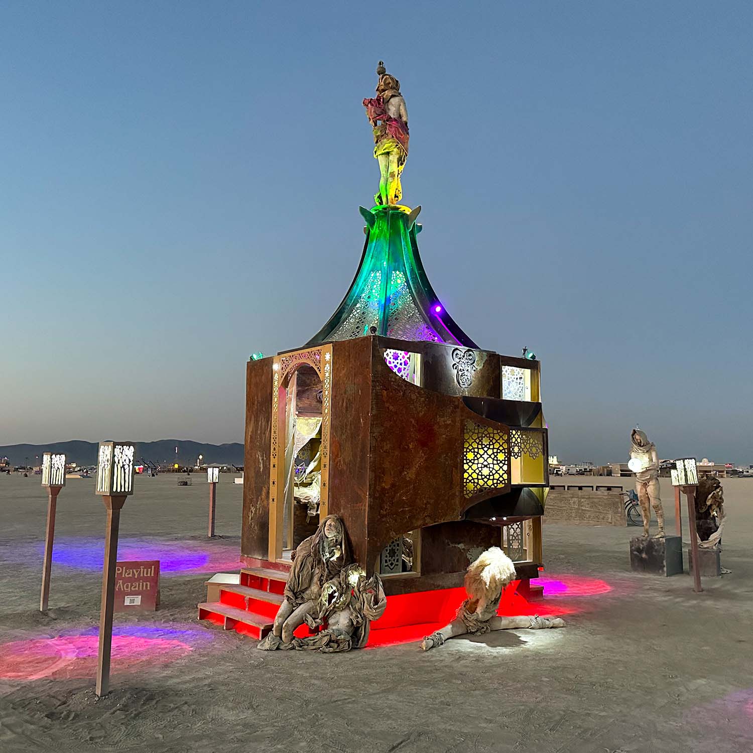 The illuminated pavilion at dusk with its red steps, the project sign and sculptures arranged around it