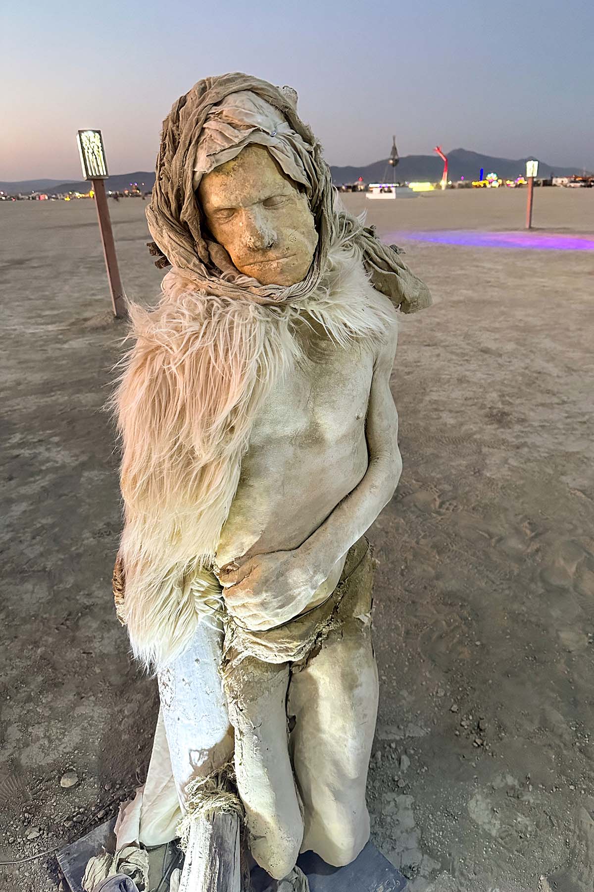 Close-up of a pale, fur-wrapped figure with a cloth hood standing on the playa at dusk