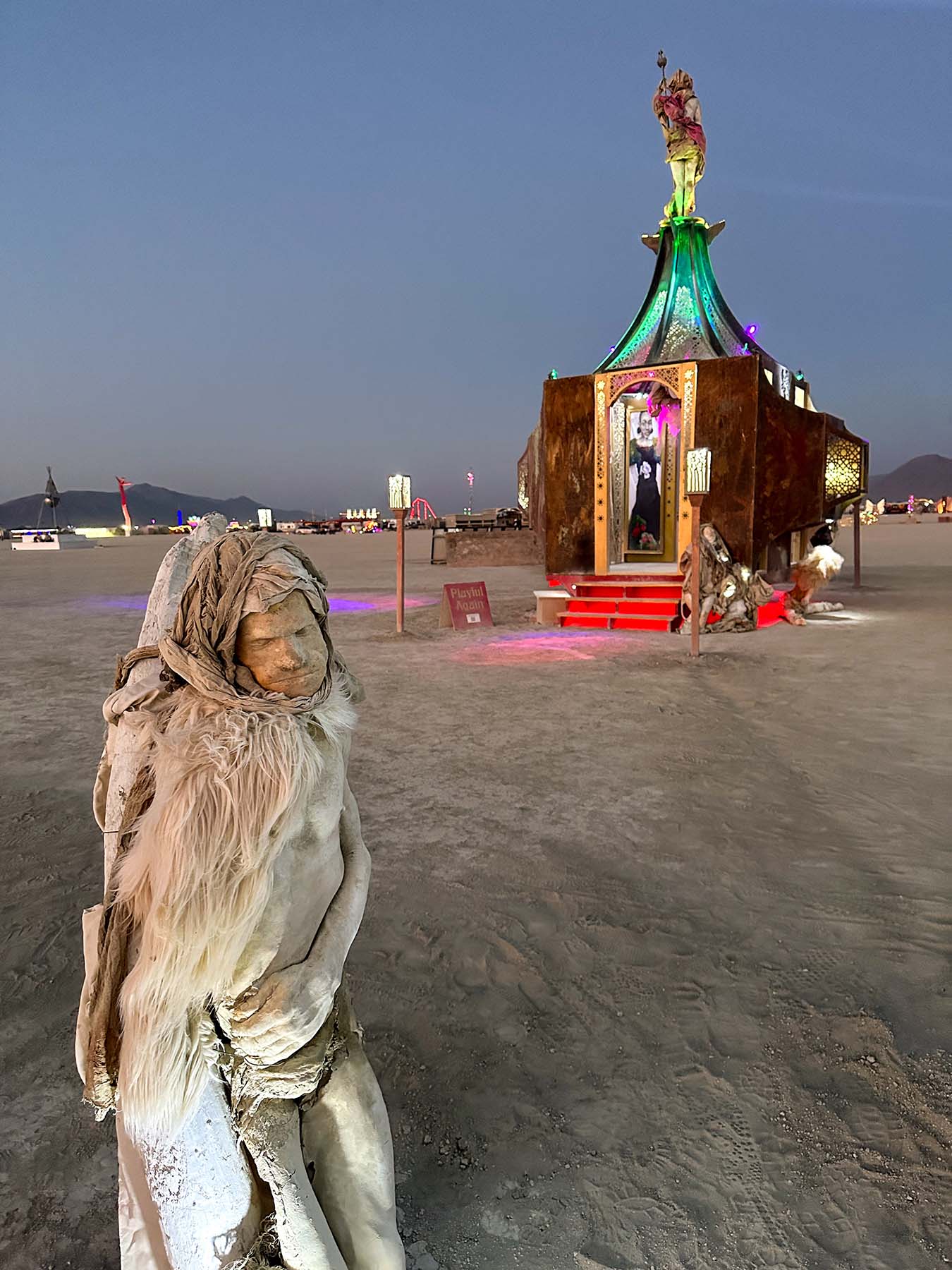 A fur-wrapped figure in the foreground with the glowing pavilion behind it at dusk