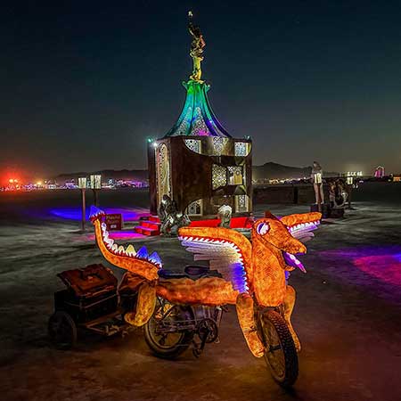An illuminated dragon-shaped art car parked beside Playful Again at night