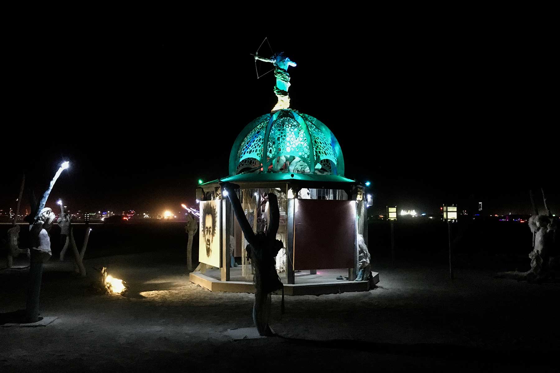 Secretly Abandoned Spaces at night: the domed structure glowing green on the playa, topped by an archer figure and surrounded by cast figures