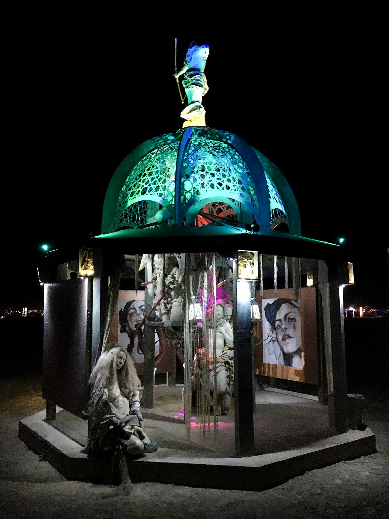 The structure at night with its teal-lit lattice dome, a long-haired cast figure seated on the front step and portrait paintings inside