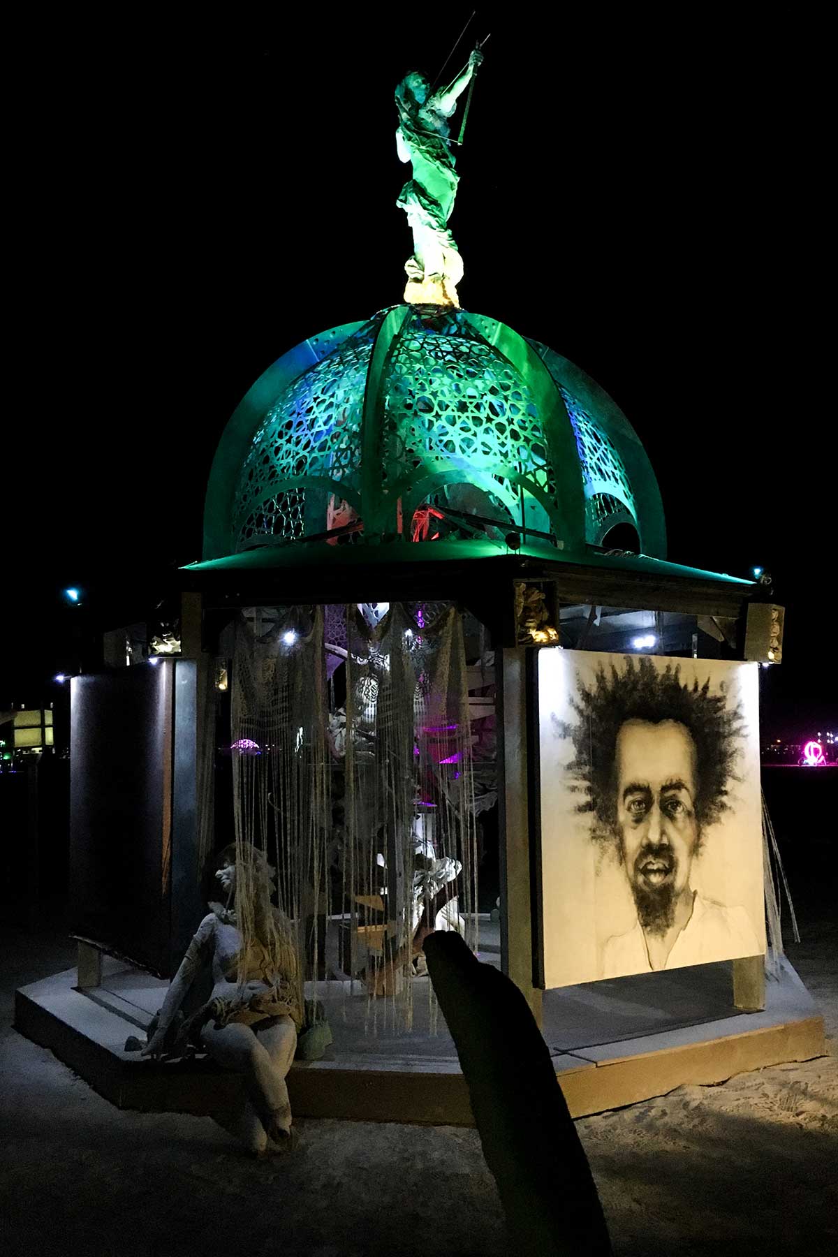 The structure at night with a large charcoal portrait of a man with wild hair on a side panel and a figure seated on the step