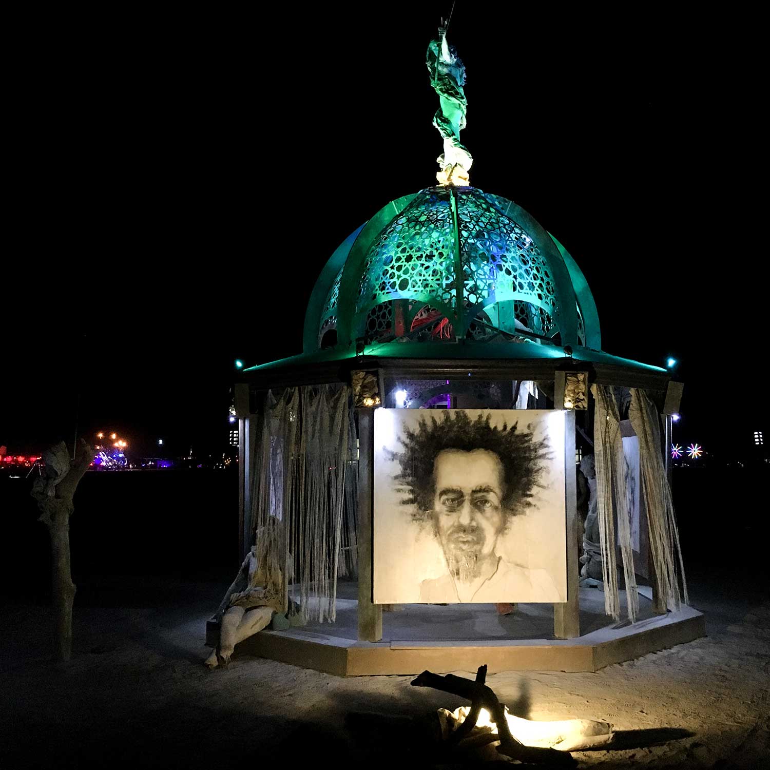 The structure at night with the charcoal portrait panel lit from below and bare branches in the foreground