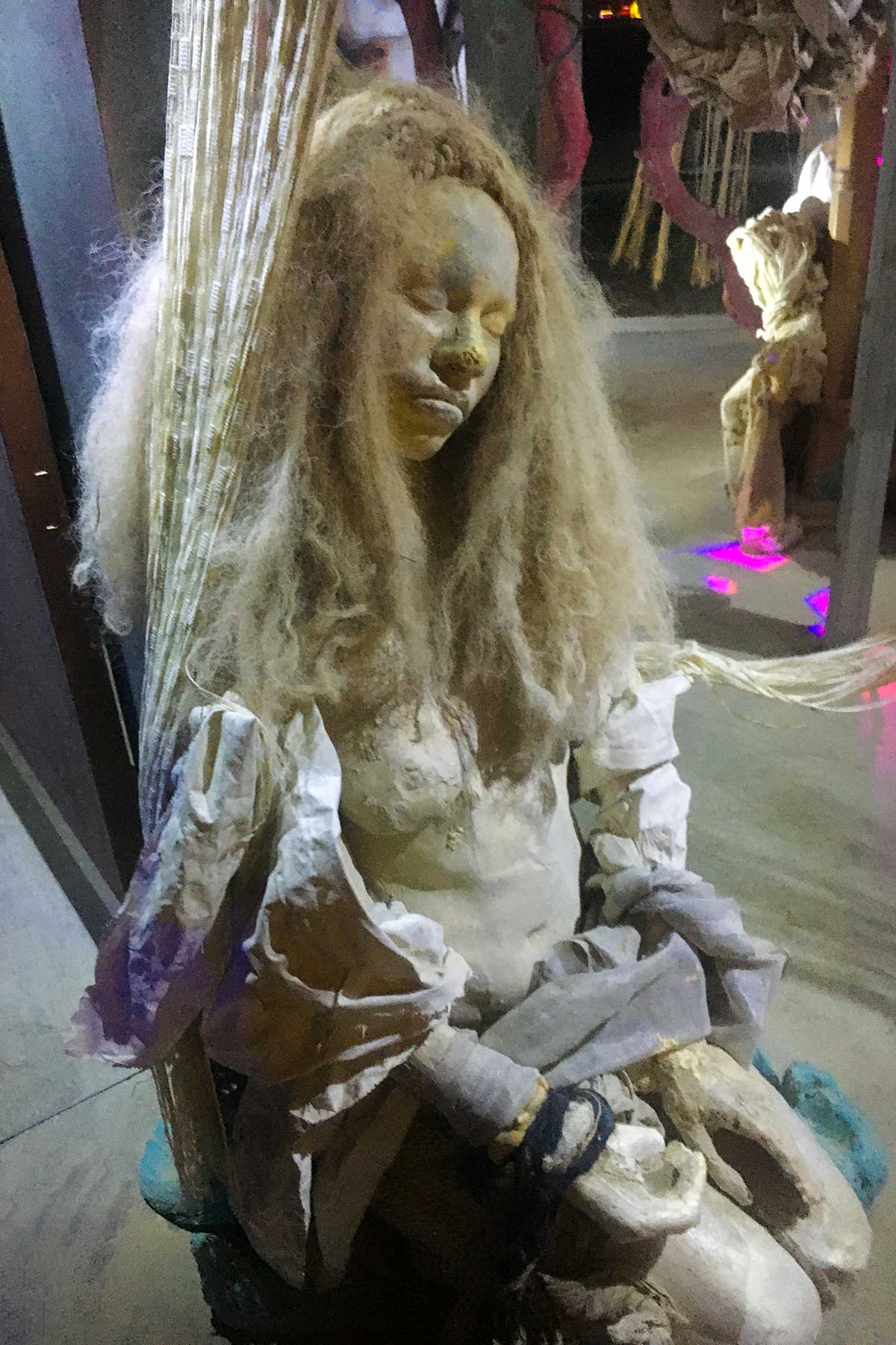 Close-up of a cast figure with long pale hair and closed eyes, seated with her hands in her lap inside the structure