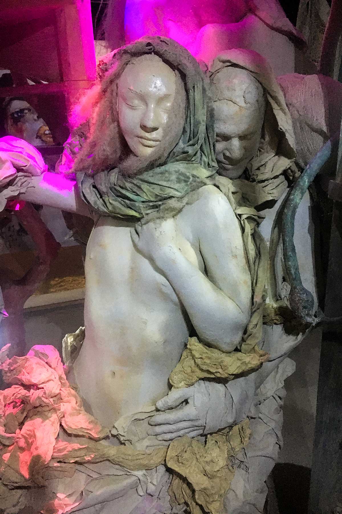 Close-up of the embracing pair at night: a hooded woman with closed eyes held from behind by a second figure, lit in magenta