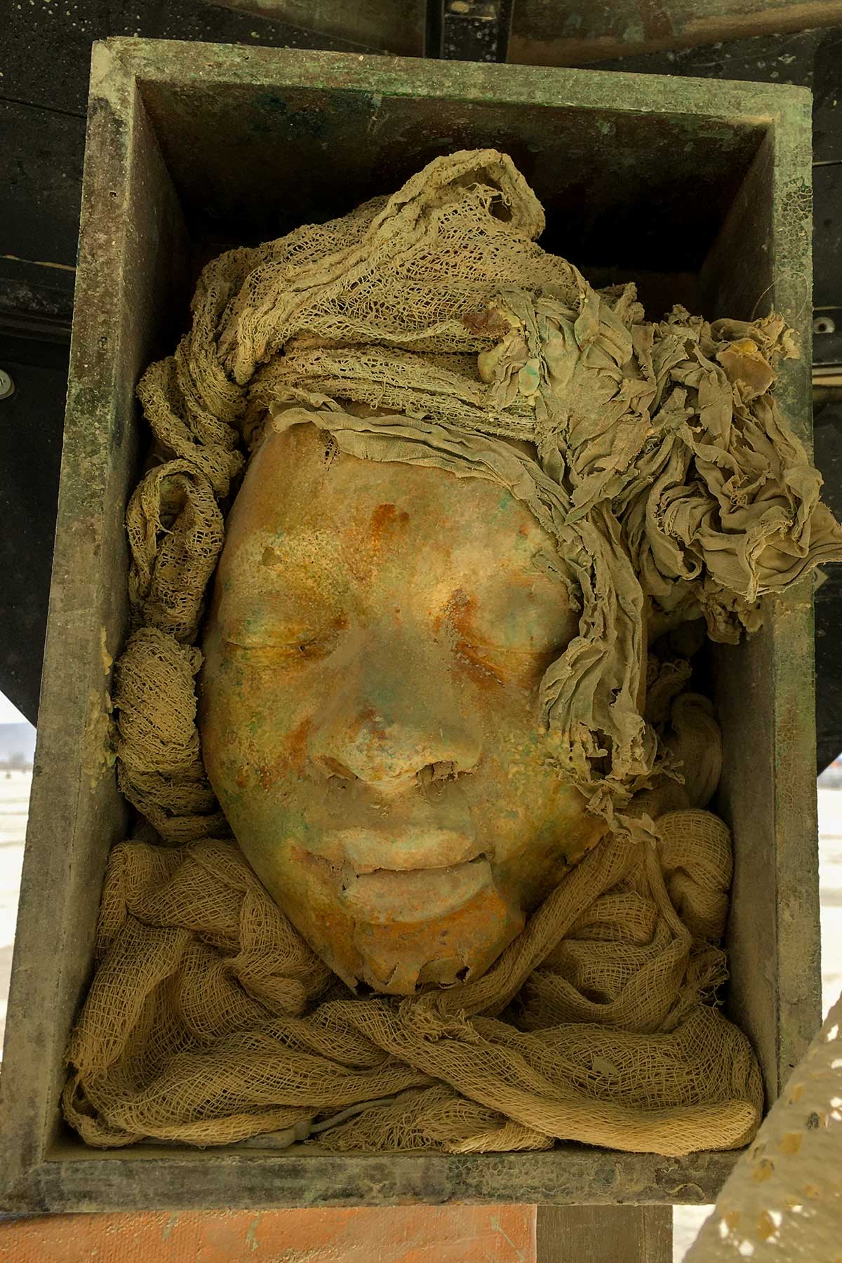 A cast face with closed eyes, framed by bunched gauze in a weathered metal box