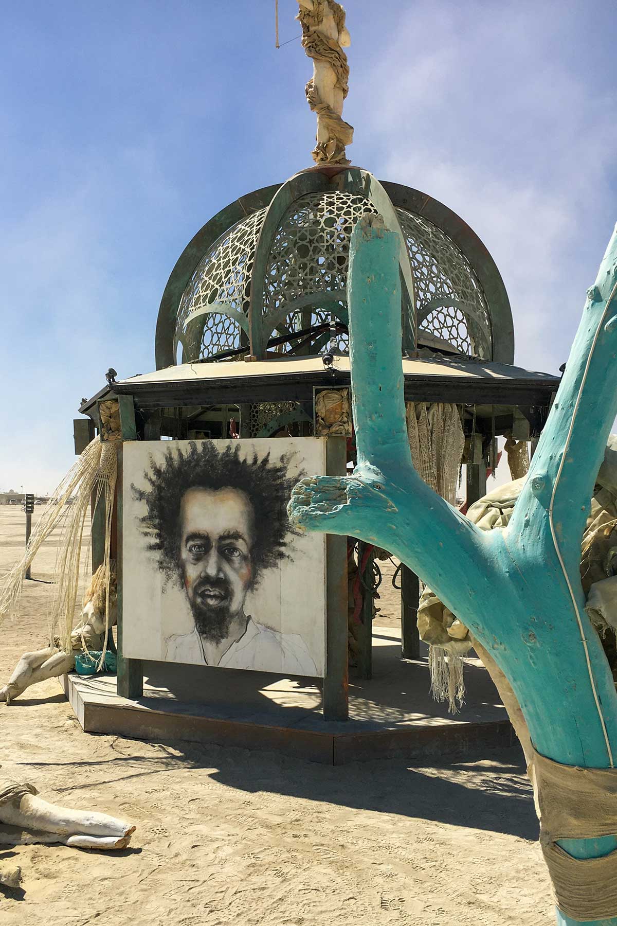The charcoal portrait panel on the structure by day, behind the branches of a turquoise tree