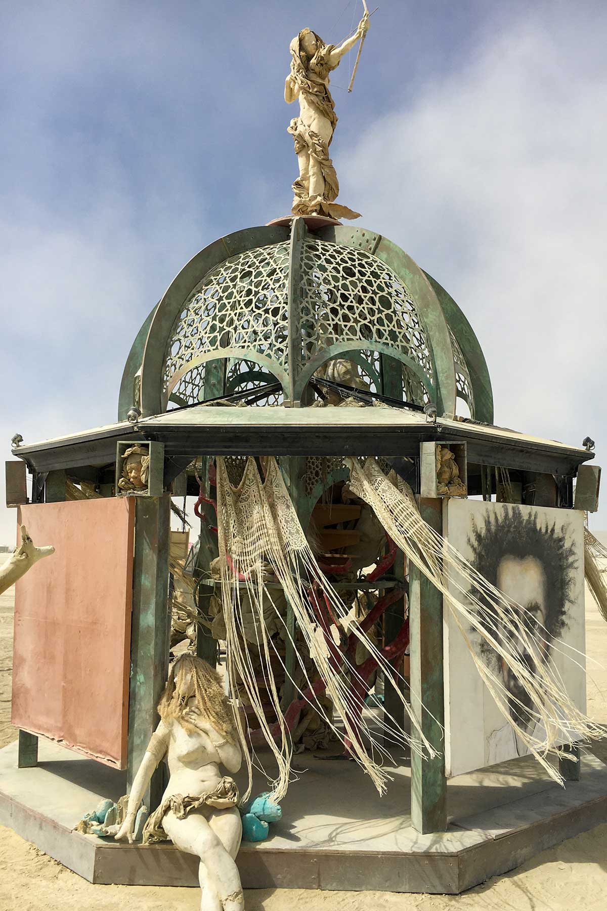 The structure by day with ragged curtains blowing from the dome, a long-haired figure seated on the step and the portrait panel at right