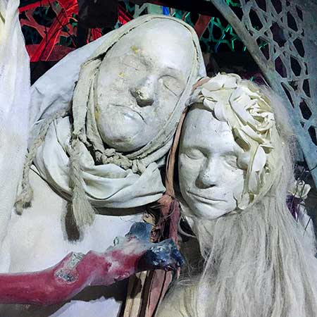 Close-up of two cast faces of a husband and wife, one hooded, the other crowned with white flowers