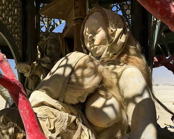 Close-up of a hooded cast mother cradling a long-haired figure, dappled by the shadow of the lattice dome