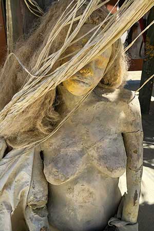 Close-up of a cracked, dust-covered cast figure with long hair, her face half hidden by hanging strands of cord