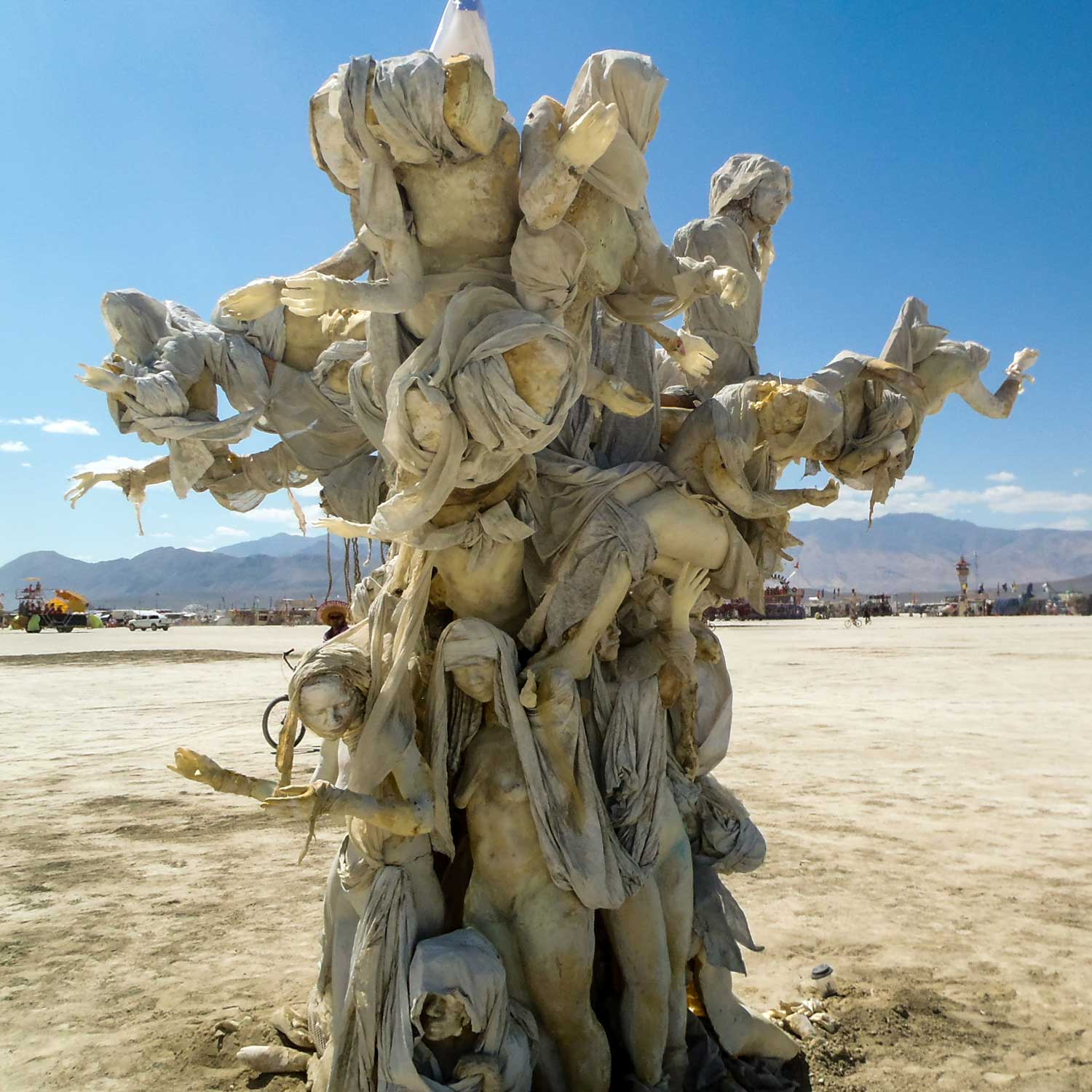 Womantree by day under a bright blue sky, with mountains and the camps of Black Rock City in the distance