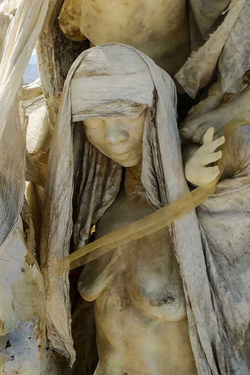 Close-up of a hooded cast figure with downcast eyes and a band of fabric across her body