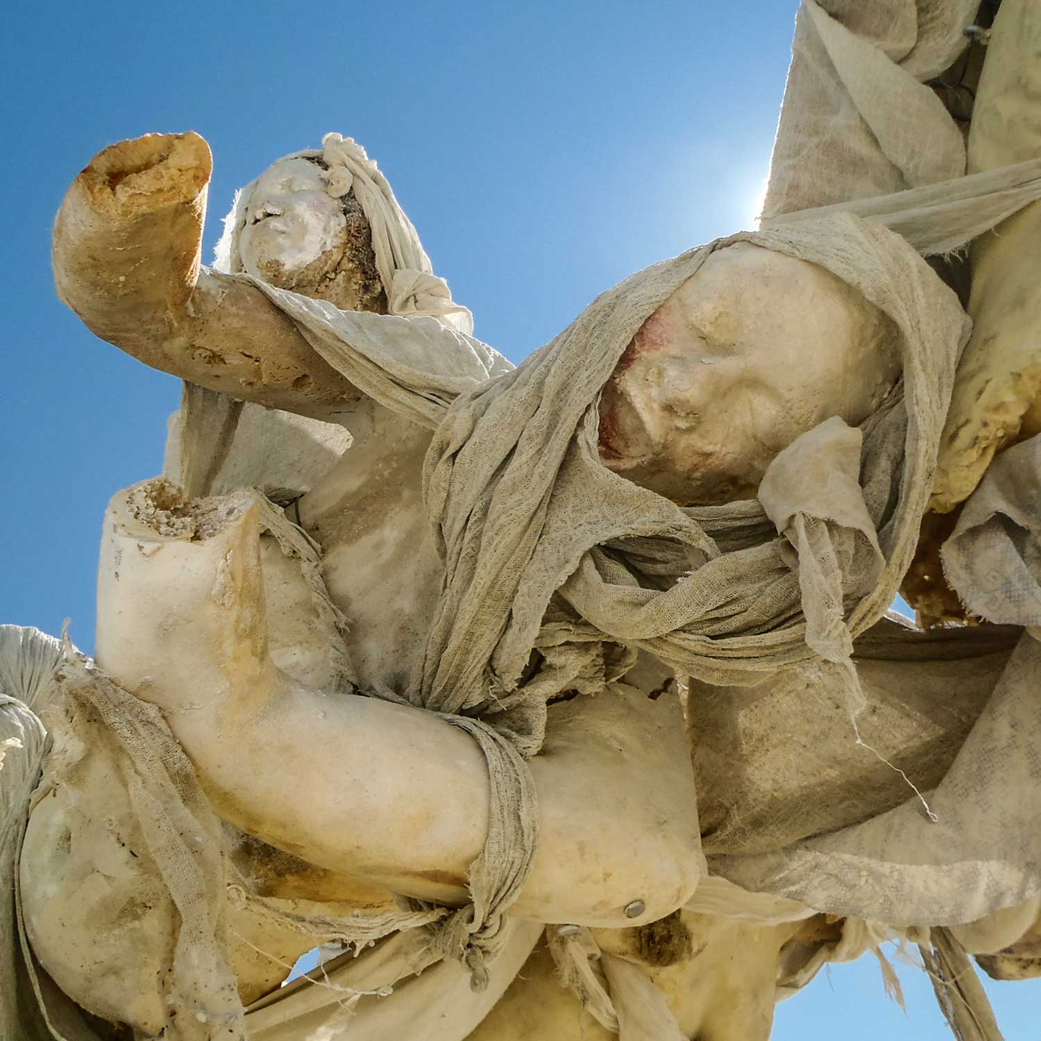 Looking up at cast figures with veiled heads and outstretched arms against a sunlit blue sky