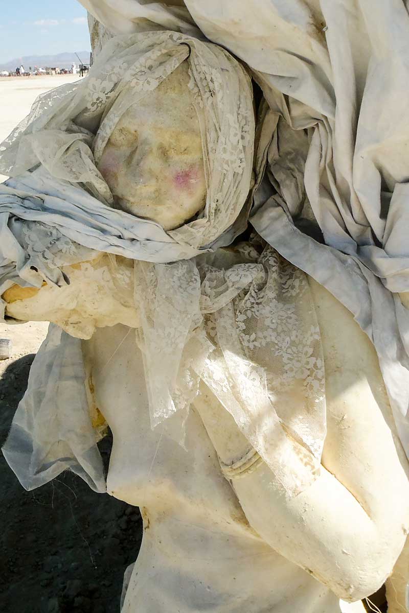 Close-up of a veiled cast figure with rosy cheeks, wrapped in sheer fabric, with the playa behind
