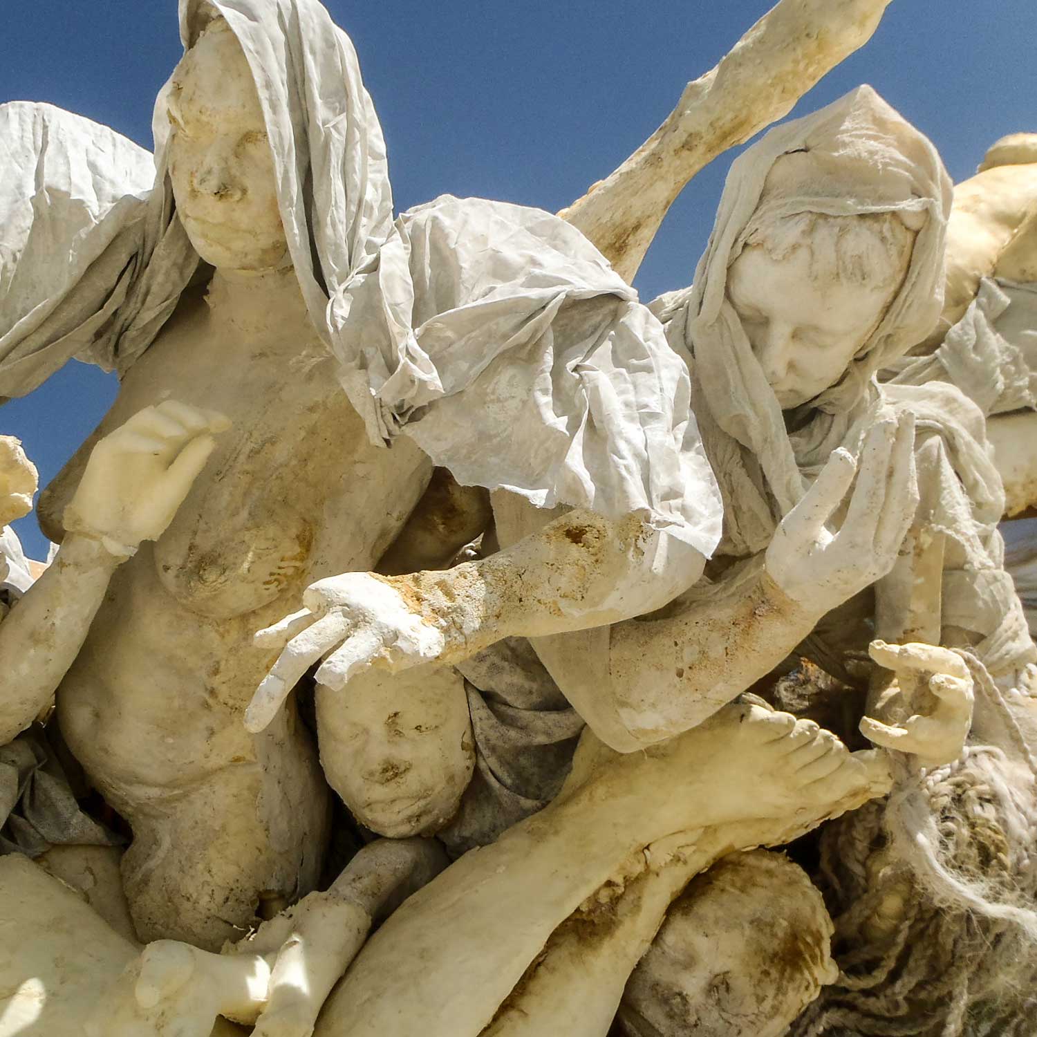 Cluster of cast figures with reaching hands and flowing fabric wraps against a blue sky