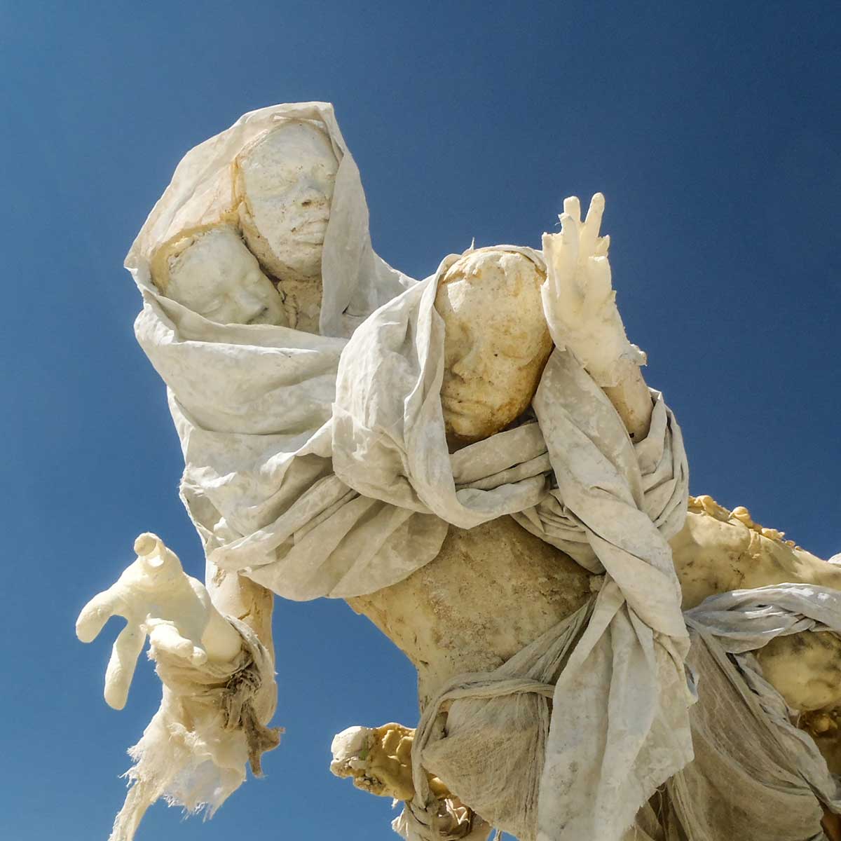 Close-up of a shrouded cast figure reaching upward with an open hand against a deep blue sky
