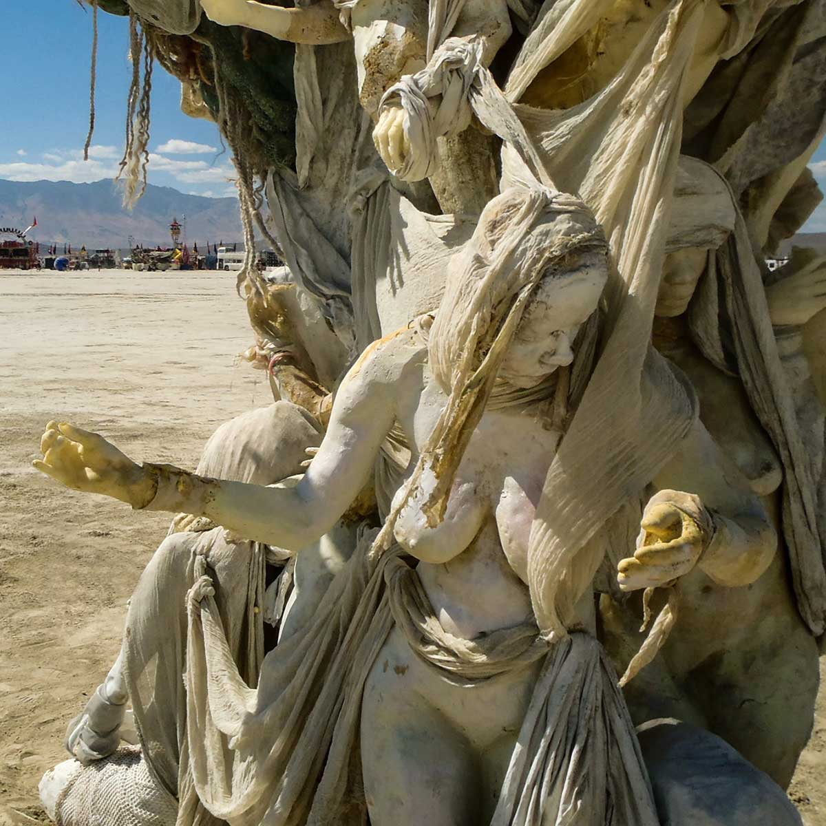 Close view of a cast woman with long draped hair reaching outward from the trunk of Womantree, with the playa and mountains beyond