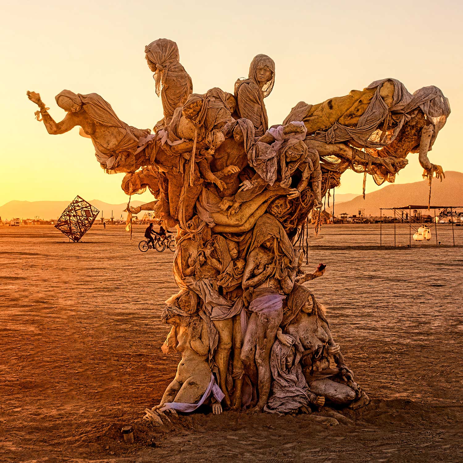 Womantree at sunset, its branches of draped cast figures glowing gold against an orange sky, with a cyclist and a geometric art piece on the playa behind