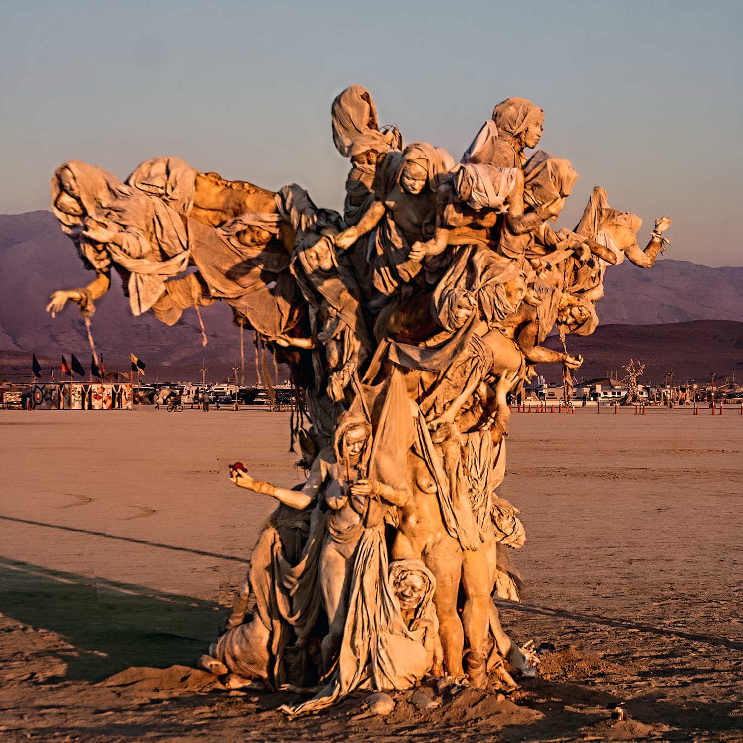 Womantree glowing orange in low sunlight, a lone figure reaching out from its trunk as long shadows stretch across the playa