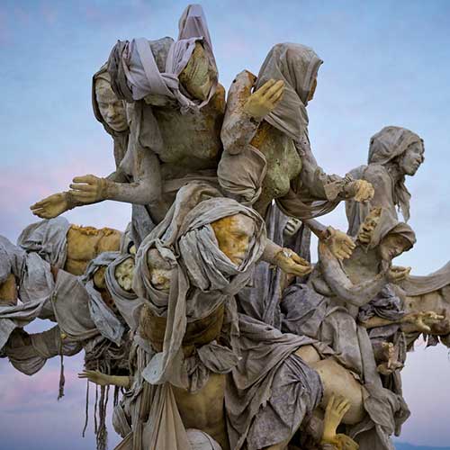 Close-up of hooded cast figures reaching outward from the crown of Womantree against a twilight sky