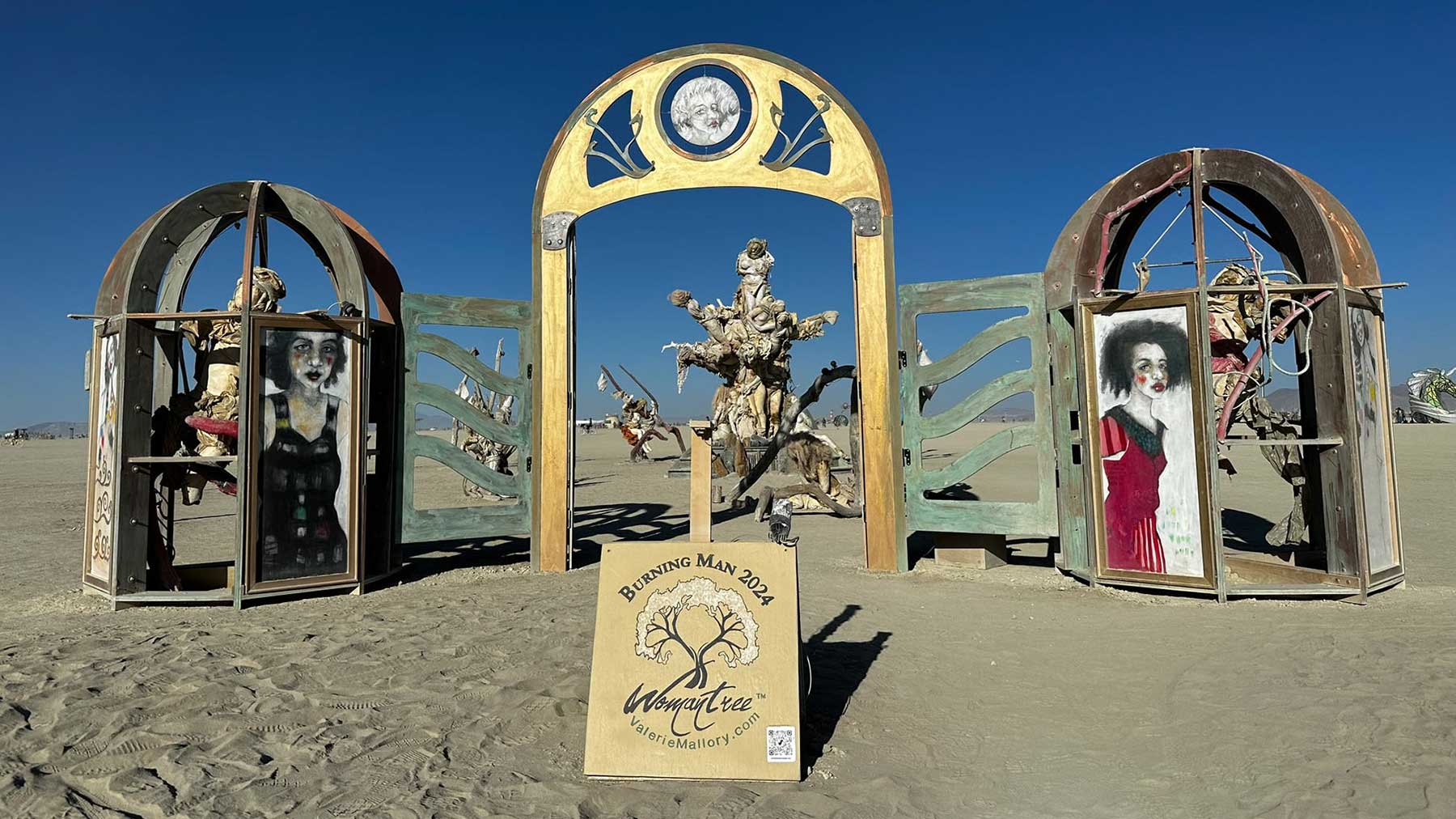 Entrance to Womantree by day: a golden arched gateway flanked by two domed portrait kiosks, with a Womantree sign in front and the tree beyond