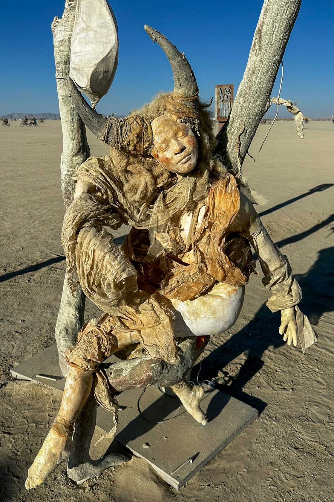 Cast figure of a horned woman draped in tattered fabric, seated on a swing on the playa