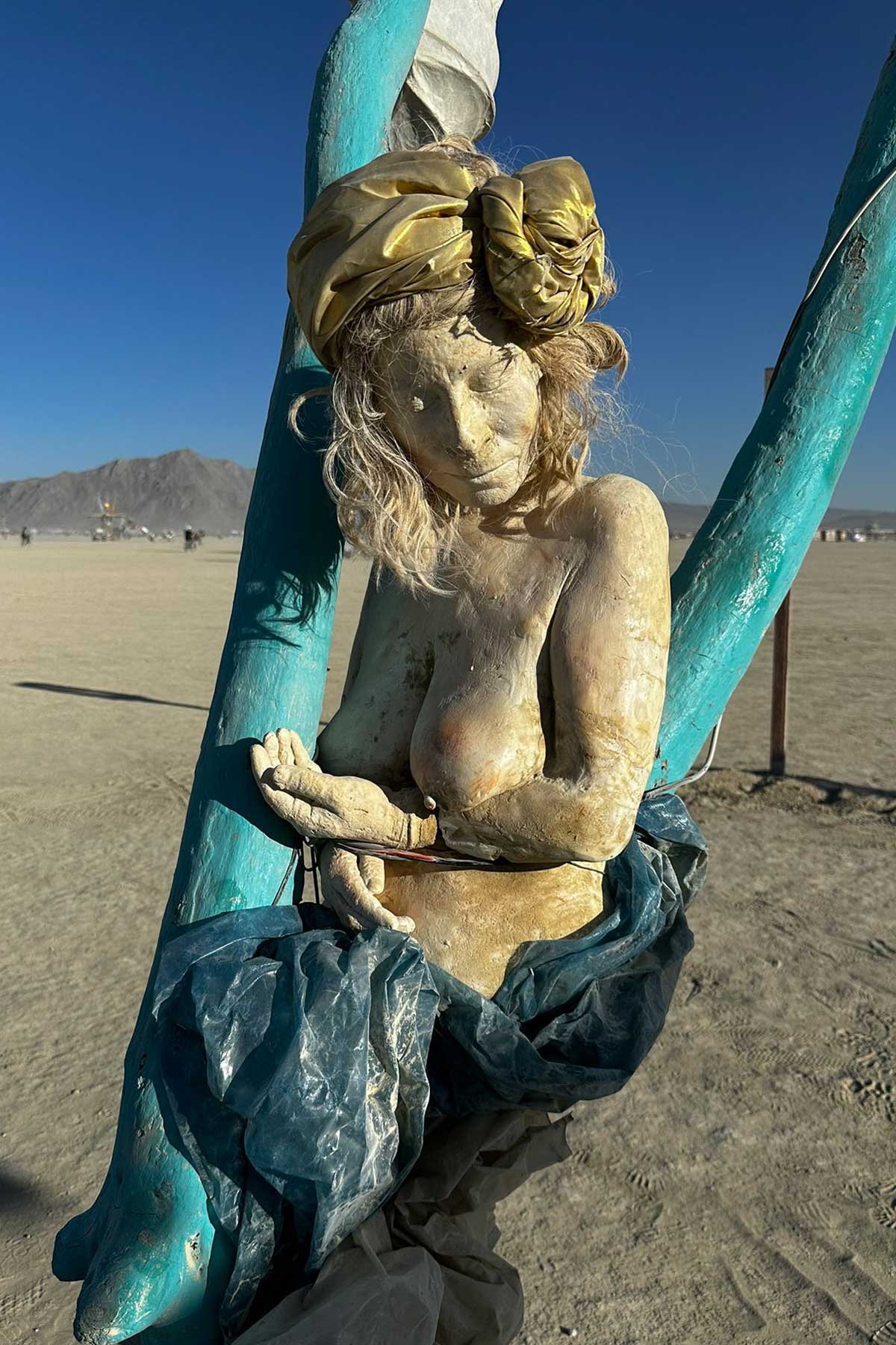 Cast figure of a woman in a golden head wrap, hands cupped together, on a teal swing