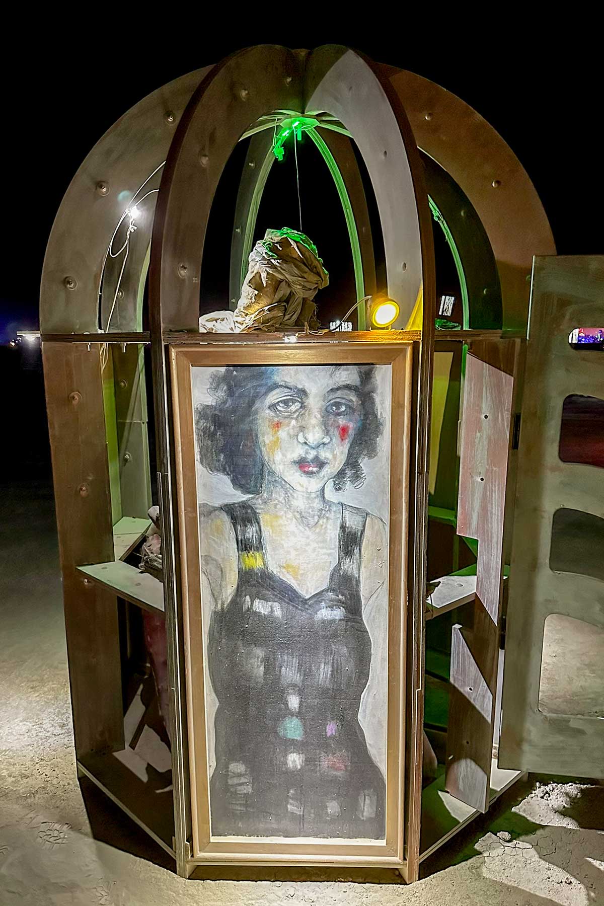 Domed kiosk lit from inside at night, its door showing a painted portrait of a woman in a patterned dress
