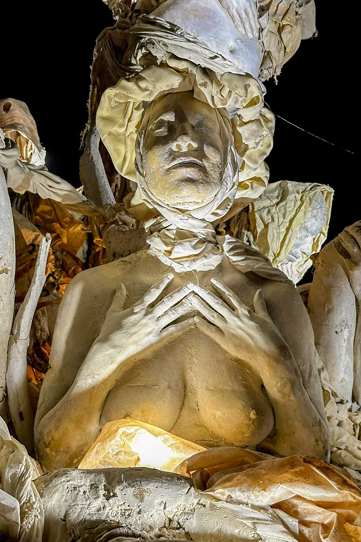Close-up at night of a cast woman with eyes closed and hands folded on her chest, lit warmly from below