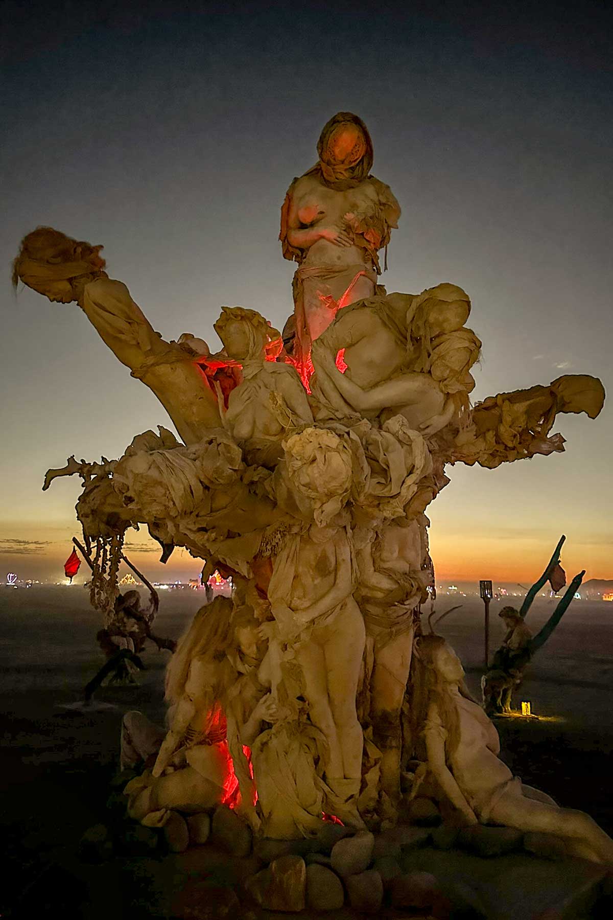 Womantree at sunset, red lights glowing among the cast figures against an orange horizon
