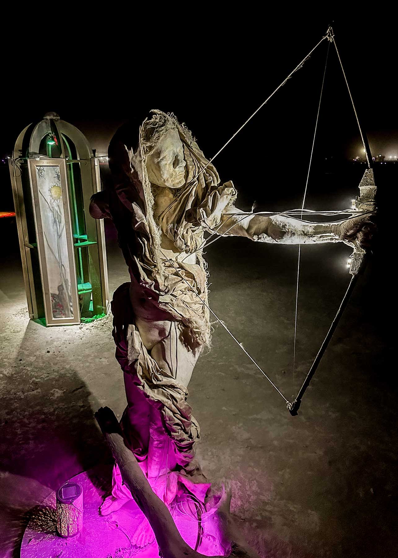 Cast figure of a woman archer drawing a bow at night, a lit portrait kiosk behind her