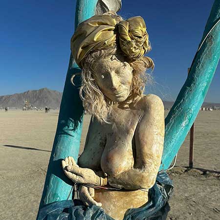 Cast figure of a woman in a golden head wrap, hands cupped together, on a teal swing