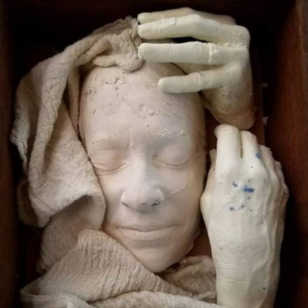 Plaster cast of a woman's face with eyes closed, framed by draped cloth and cast hands