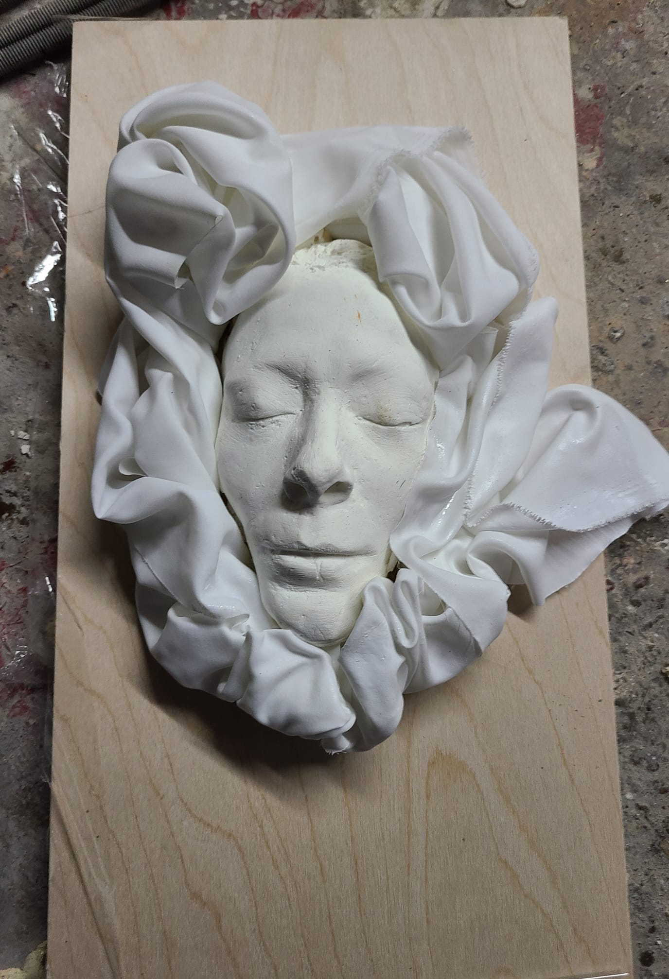 White plaster face cast surrounded by sculpted fabric folds, mounted on a wooden board
