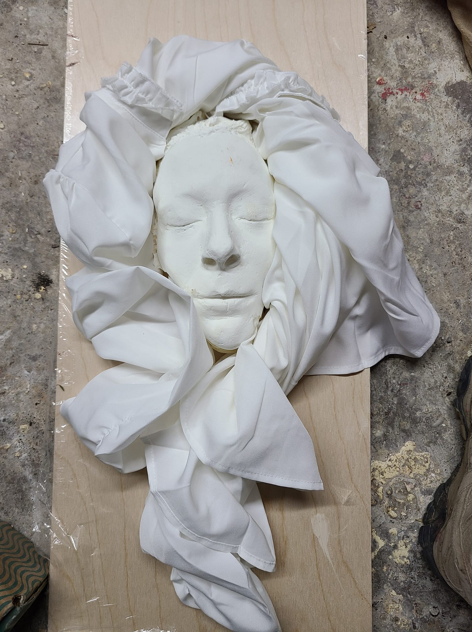 White face cast with eyes closed, wrapped in flowing plaster-stiffened fabric on a wooden board