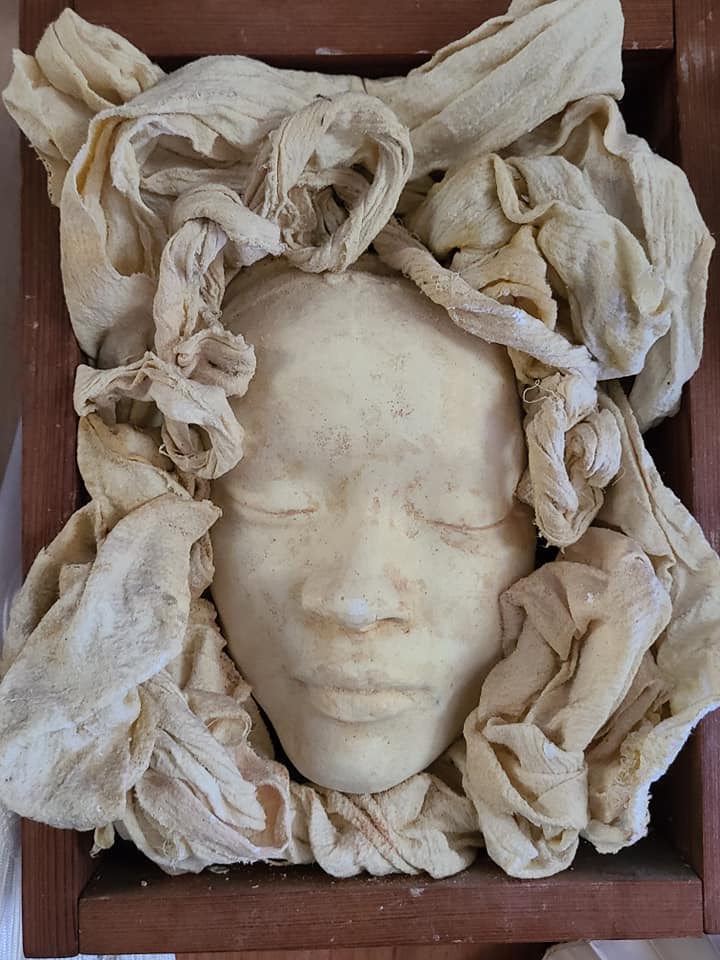 Face cast with eyes closed, set in a wooden box among ruffled, plaster-stiffened cloth