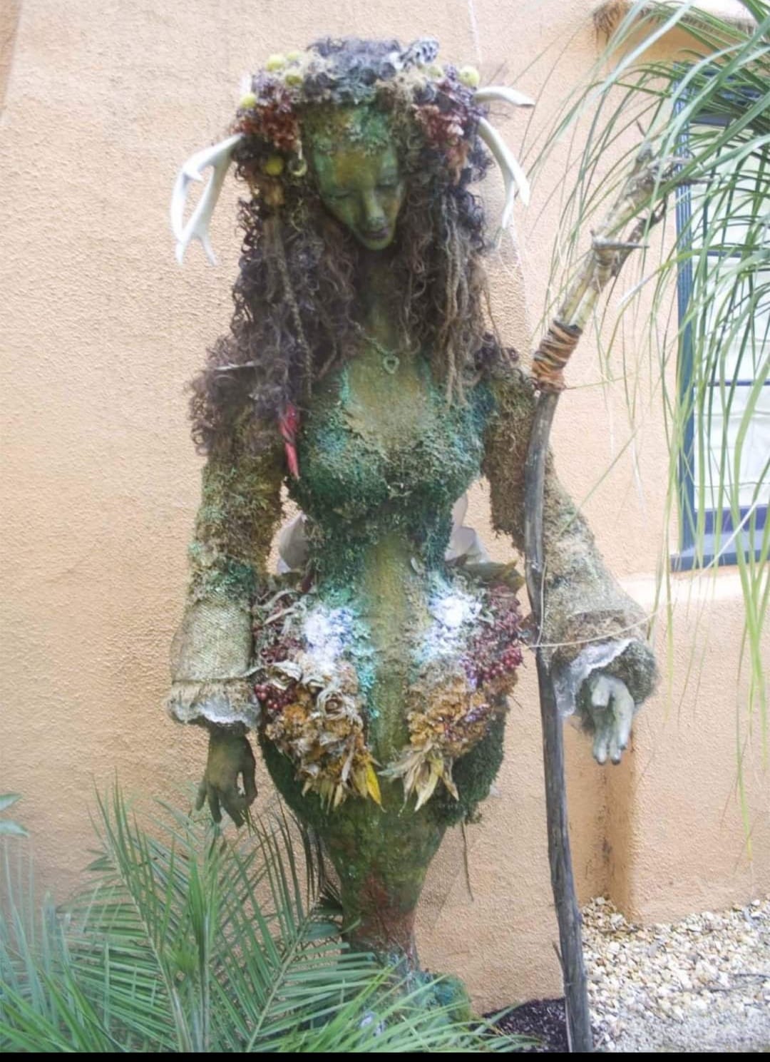 Standing figure covered in moss, dried flowers and greenery, wearing a flowered crown