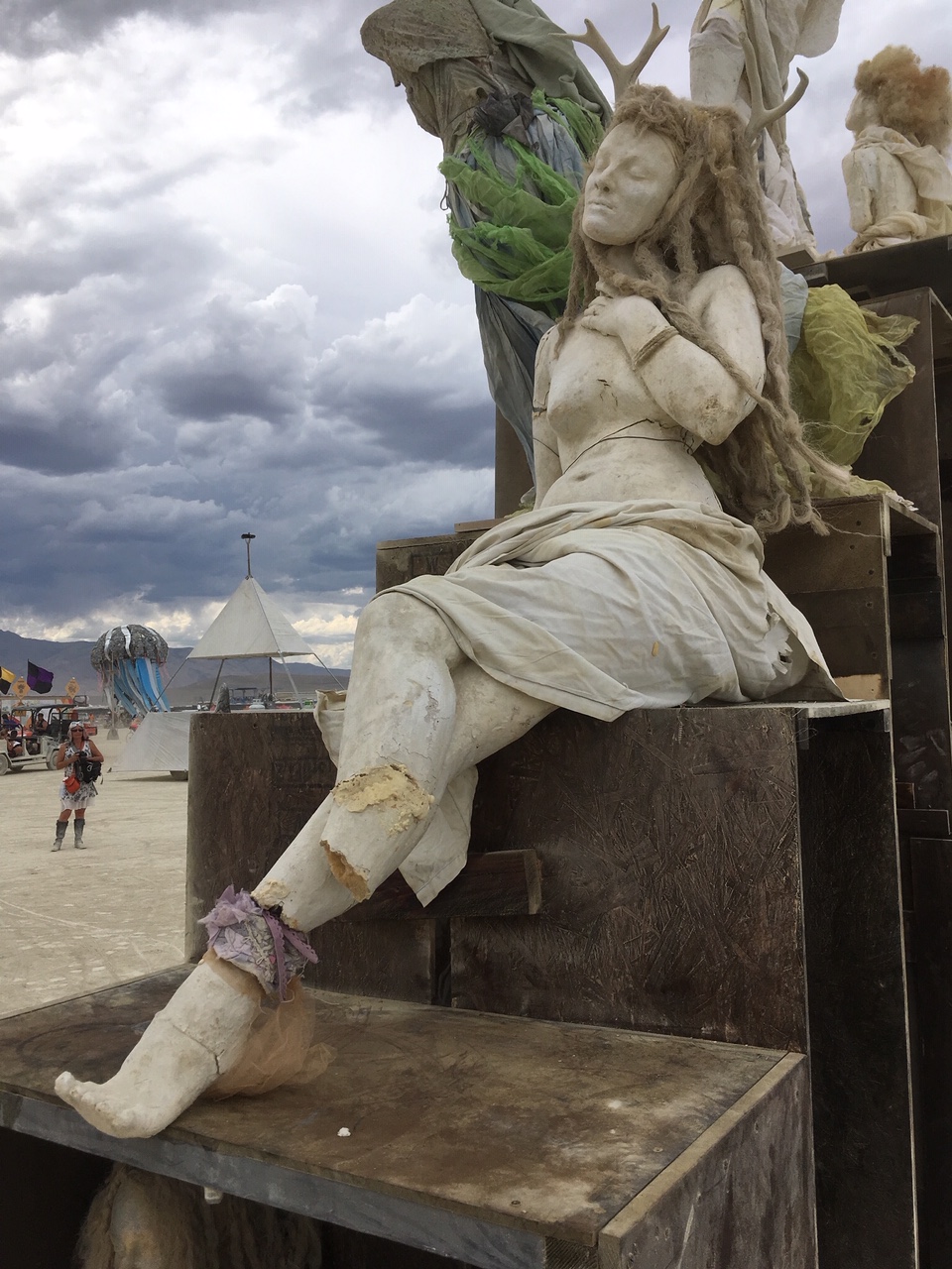 Seated cast figure in draped cloth perched on wooden crates on the playa under stormy skies